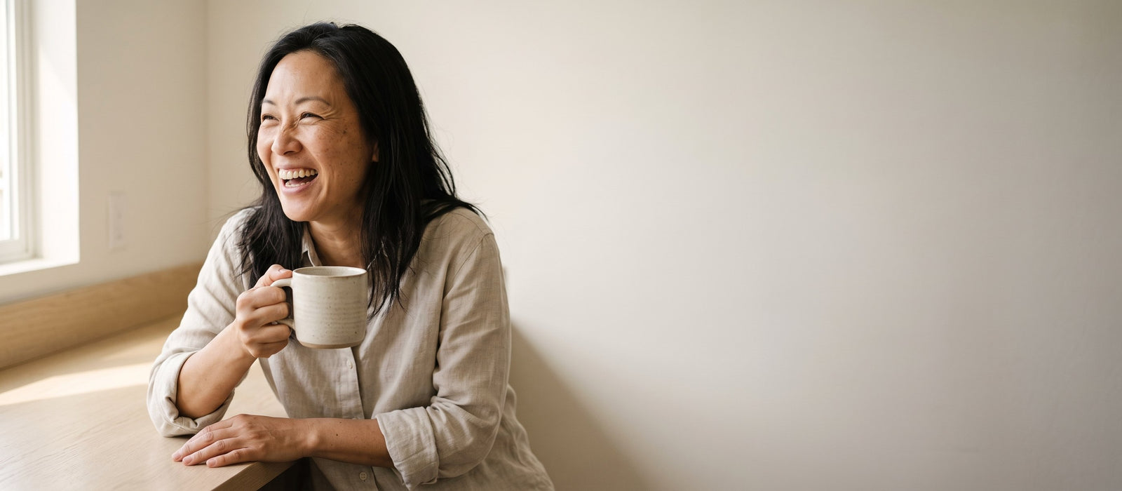 A woman laughing at her kitchen counter in the morning, a mug of coffee in her hand